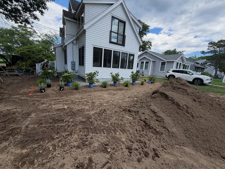 Plants being arranged in prepared garden beds beside a white home.